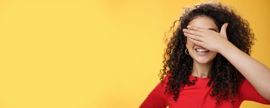 Lifestyle. Close-up shot of dreamy happy young cute woman with curly hair covering eyes with palm as counting or playing peekaboo smiling broadly anticipating surprise happen over yellow background.