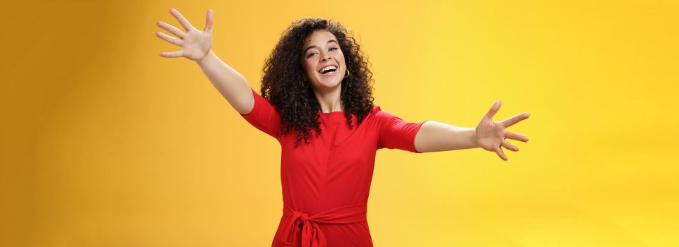 Come into my arms. Portrait of friendly and loving, caring charming woman with curly hair in red casual dress spread hands as wanting give hug smiling broadly at camera giving warm welcome or cuddle.
