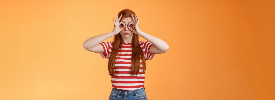 Cute silly redhead charming girl making finger glasses, look through okay circle signs amused, pouting childish, acting silly, searching something, looking for sales, stand orange background.