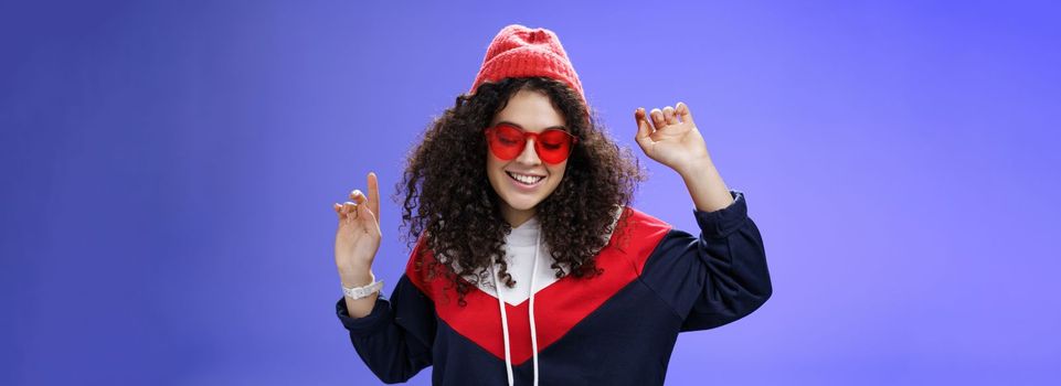 Girl feeling awesome having fun and enoying cool party, dancing with raised hands looking down smiling broadly wearing sunglasses and stylish beanie posing against blue background.