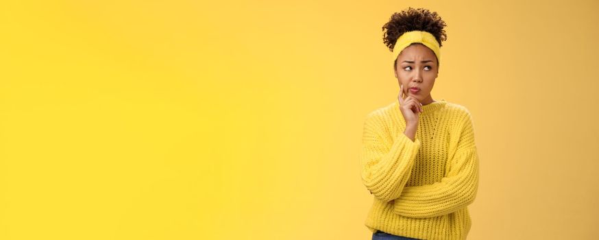 Perplexed unsure cute timid african-american female thinking how escape awkward situation smirking frowning concerned touch cheek thoughtful thinking nervously, worried yellow background.