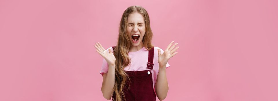 Young girl scared to death seeing spider in room yelling out loud closing eyes and opening mouth gesturing with raised hands near chest feeling afraid and insecure posing against pink background. Copy space