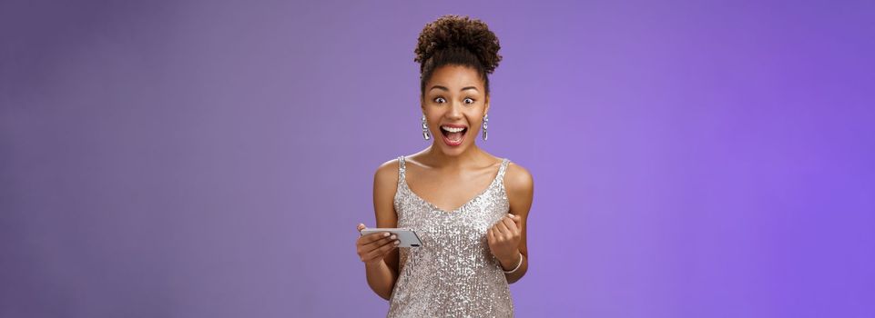 Excited charming young african-american woman lucky winning game playing smartphone standing pleased yelling happily clench fist triumphing joyfully celebrating great news received message.