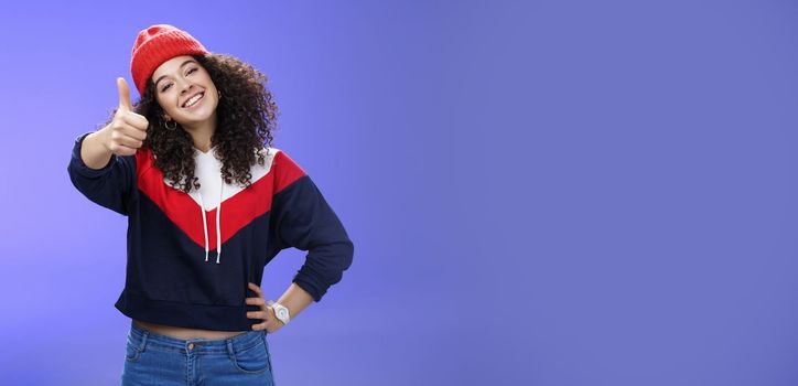 You doing great, like. Portrait of happy delighted young stylish woman in winter beanie and sweatshirt showing thumbs up pleased and satisfied approving and being supportive smiling at camera.