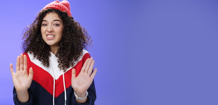 Lifestyle. Displeased european woman with curly hair raising palms near chest and waving in refusal and rejection gesture grimacing unsatisfied and unwilling to participate in suspicious action over blue wall.
