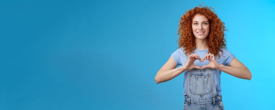 Tenderness, love, romance concept. Lovely feminine cute redhead curly woman show heart gesture chest express sympathy passionate feelings smiling broadly cherish feelings standing blue background.