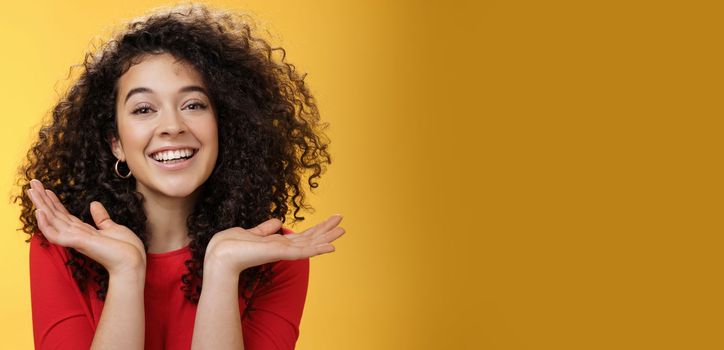 Lifestyle. Close-up shot of happy kind and tender pretty caucasian female student with curly hair and perfect skin smiling delighted holding palms spread near face having fun over yellow background.