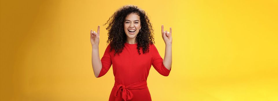 Rock n roll live. Charismatic excited young curly-haired woman in red dress feeling awesome as waiting concert of favorite band showing rock gestures with raised hands and smiling thrilled and happy.