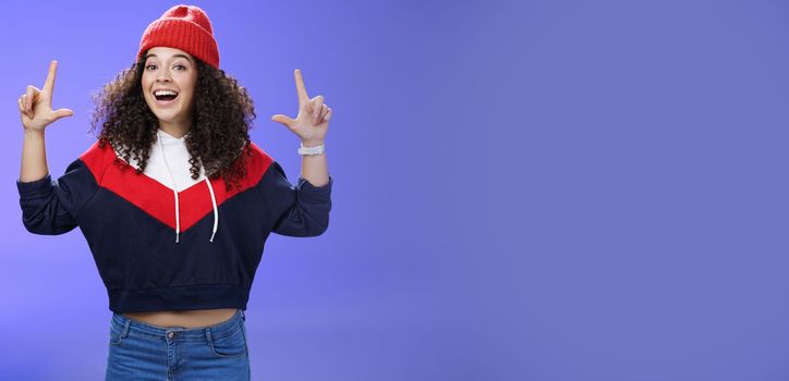 Lifestyle. Waist-up shot of cheerful energized young woman calling friend inviting hang out as weather awesome pointing up with raised hands smiling broadly posing in red winter hat over blue background.