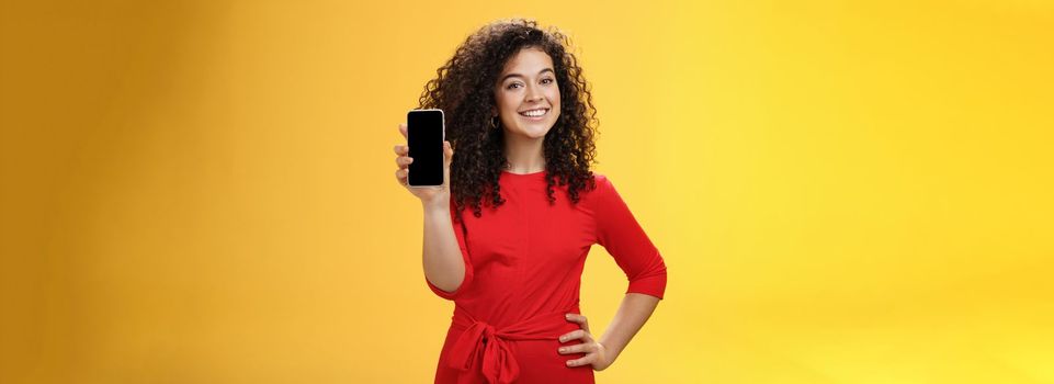 Girl brag with new phone she got on christmas feeling delighted holding mobile device in hand showing smartphone screena t camera, smiling broadly with uplifted mood over yellow background. Technology concept