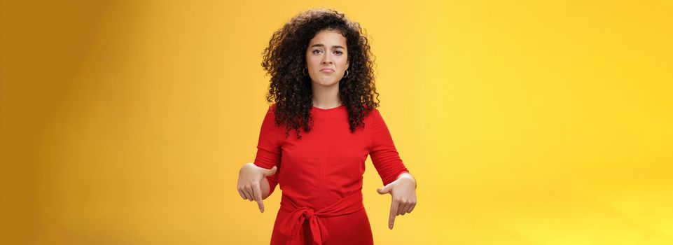 Portrait of gloomy girl with sad face pursing lips down frowning disappointed and pointing down with regret and unsatisfied expression, missing opportunity feeling upset over yellow background.