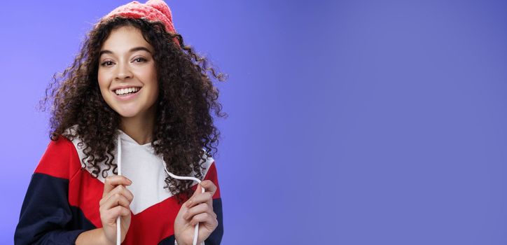 Close-up shot of carefree attractive feminine girl with curly hair in beanie playing with sweatshirt as posing over blue background, smiling at camera having fun and positive attitude.