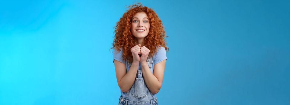 Pretty please buy me that. Amused longing excited redhead cheerful girl clench fists together stare camera desire express aspiration wanna receive buy product standing blue background thrilled.