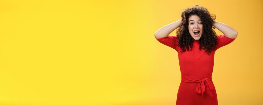 Girl feeling pressure standing anxious in panic holding hands on curly hair yelling at camera disturbed, freaked out being tensed and upset with bad situation, standing troubled over yellow background.