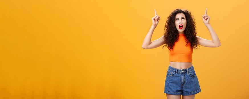 Silly and moody european party girl with curly hairstyle. in red lipstic and stylish orange cropped top complaining on bad weather raising hands pointing and looking up with displeased unhappy face.