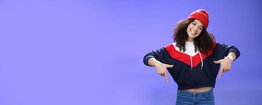 Lifestyle. Waist-up shot of cool and stylish young woman with curly hair smiling flirty and joyful pointing down showing promotion as tilting head happily and posing over blue background in outdoor clothes.
