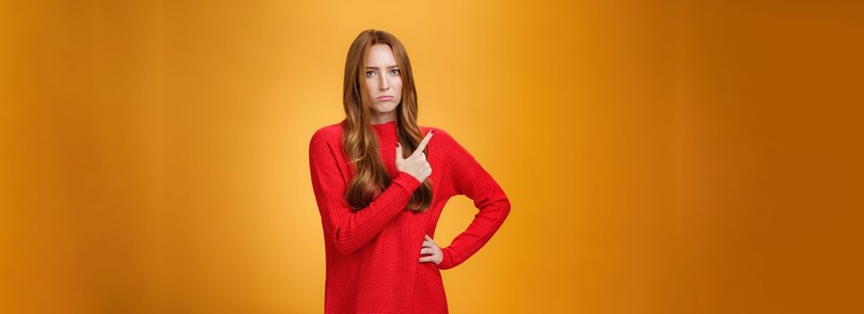 Upset and disappointed young ginger girl in red sweater pointing at upper left corner or behind frowning looking gloomy and angry, standing jealous and moody against orange background.