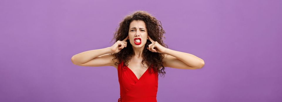 Woman fed up of hearing cursing words and arguments in her turn. Portrait of displeased insecure and offended female with curly hairstyle in red dress closing eyes and talking displeased and bothered. Copy space