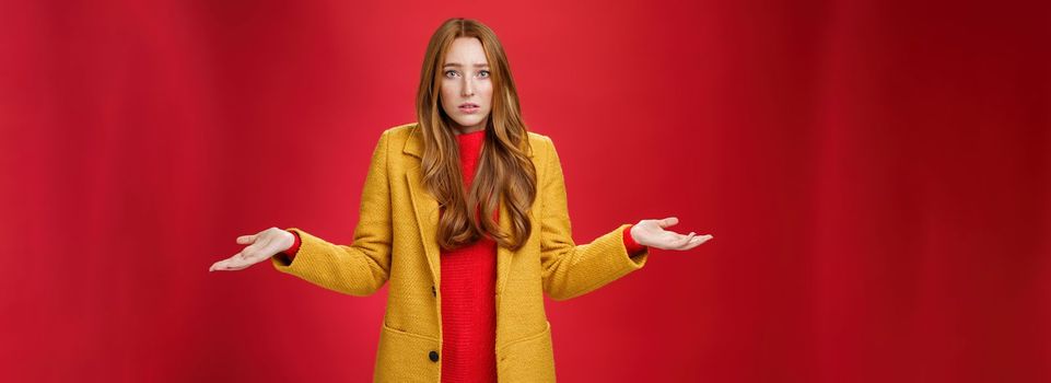 Upset and confused redhead girlfriend cannot understand what happened shrugging with spread hands sideways and clueless grimace, being unaware why, posing concerned over red wall. Body language and facial expressions concept