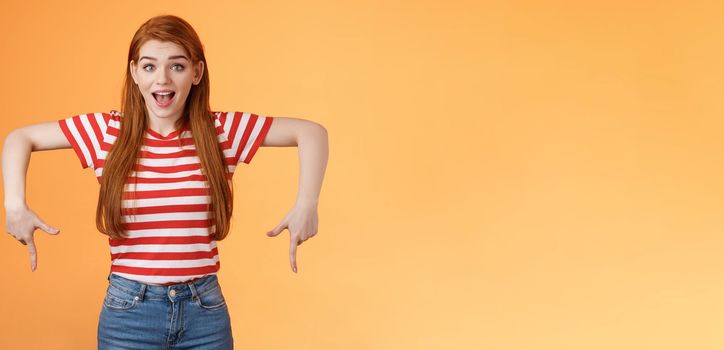 Enthusiastic lively cute redhead woman introduce incredible offer, pointing down, bottom copy space, smiling amazed, look camera, explain you awesome opportunity, feel excitement, orange background.