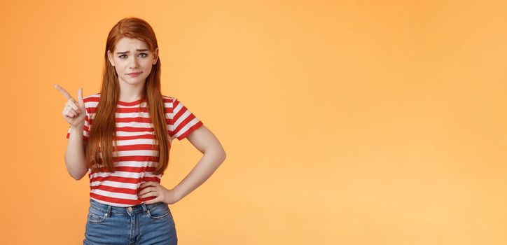 Doubtful redhead girl not buy your talks, smirking hesitant express disbelief, unimpressed about suspicious offer, pointing upper left corner wondered, stand orange background uncertain.