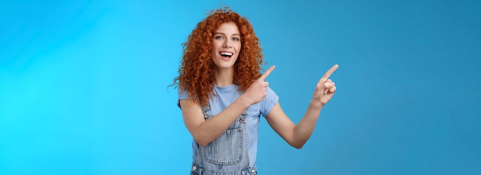 Hey check this out. Enthusiastic redhead curly stylish summer girl inviting beach party pointing upper left corner directing awesome location smiling broadly discuss awesome promo blue background.