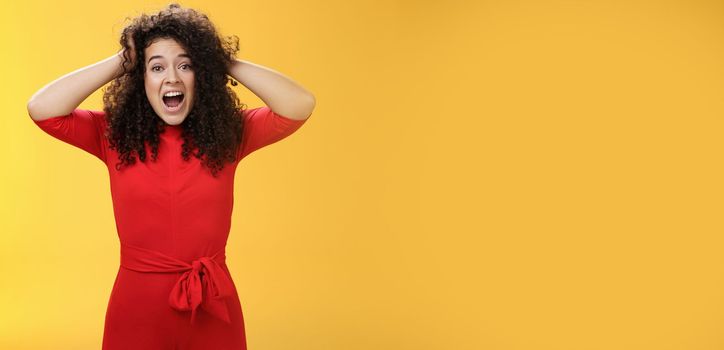 Girl feeling pressure standing anxious in panic holding hands on curly hair yelling at camera disturbed, freaked out being tensed and upset with bad situation, standing troubled over yellow background.