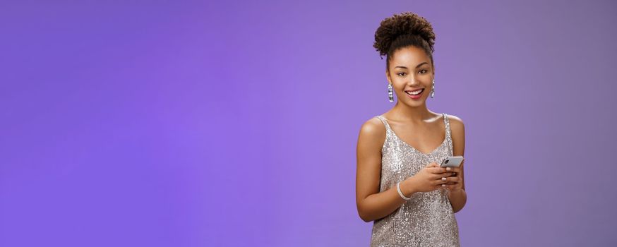 Attractive stylish confident african-american woman in silver shiny dress holding smartphone messaging scrolling internet gadget looking lucky happily camera standing blue background rejoicing.
