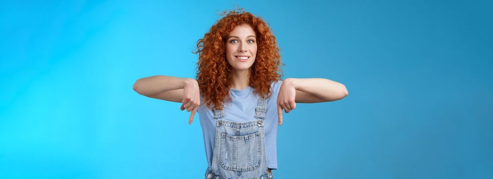 Excited happy smiling caring redhead curly-haired girlfriend thrilled awaiting summer holiday trip pointing down index finger upbeat mood show favorite store buy best prices, blue background.