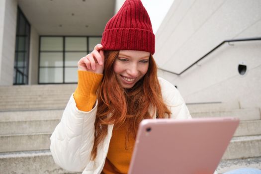 Beautiful redhead female model, ginger girl with digital tablet, sits on stairs outdoors, reads on her gadget, uses internet application and wifi on street.