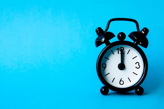 Black alarm clock isolated on blue background. The clock set at 12pm. Copy space.