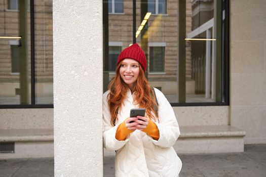 Beautiful redhead girl with smartphone, standing on street, holding mobile phone, using taxi app, connects to public wifi and uses internet, sends a text message.