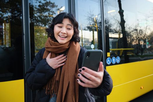 Portrait of cheerful asian girl talks on mobile phone, video chats, looks amazed at smartphone camera, stands on bus stop.