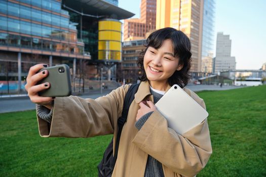 Happy university student, korean girl takes selfie with her papers and digital tablet, holds smartphone and poses near university campus.