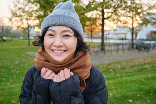 Beautiful young asian woman in warm hat and scarf, smiles at camera, walks around park on chilly spring day, posing with happy face expression.