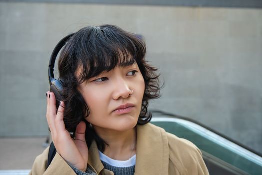 Portrait of complicated young korean girl in headphones, frowns and looks disappointed while stands and waits for public transport on street.