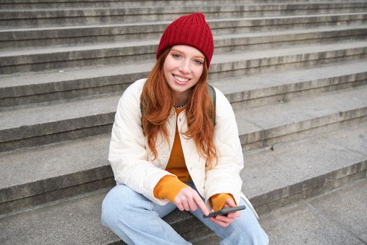 Stylish young redhead woman, talking on mobile phone app, using social media application, looking for something online on smartphone, sits on stairs outdoors.