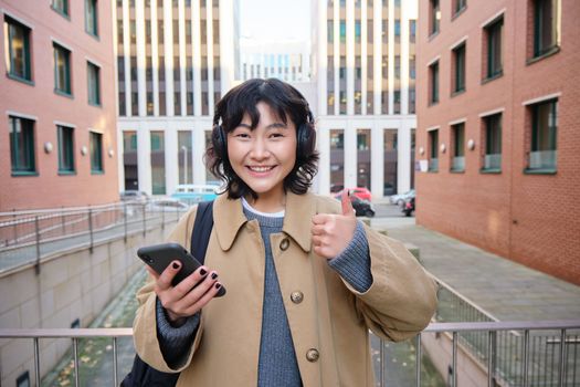 Portrait of cheerful asian girl in headphones, stands on street and shows thumbs up, found something online on her smartphone, using mobile phone.