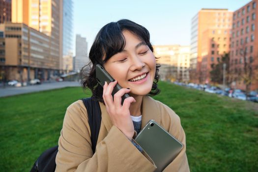 University lifestyle. Young asian student, girl with tablet and notebook, talks on mobile phone while walks from campus on street.