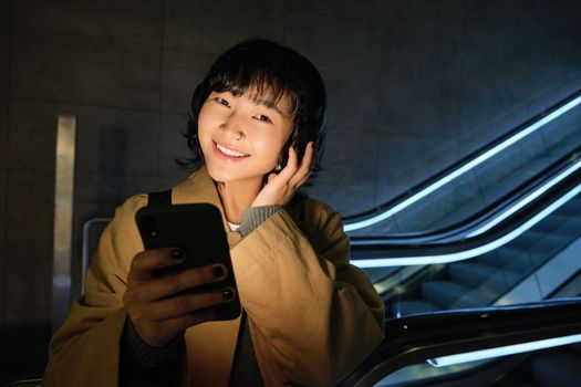 Portrait of beautiful korean girl in headphones, stands near escalator, travels, commutes home on public transport, holds smartphone, poses with mobile phone.