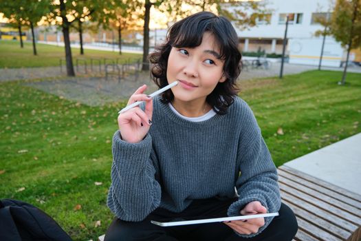 Portrait of asian woman, student in park, sits on bench with digital tablet and a pen, thinking, looking aside thoughtful, making notes, does her homework outdoors.