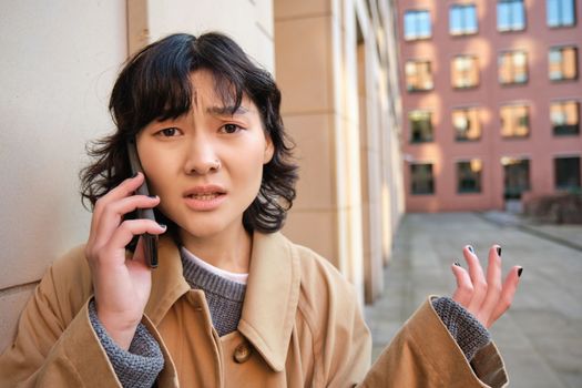 Korean girl with concerned face talks on mobile phone and shrugs, frowns and looks concerned, being lost in town, stands on street.