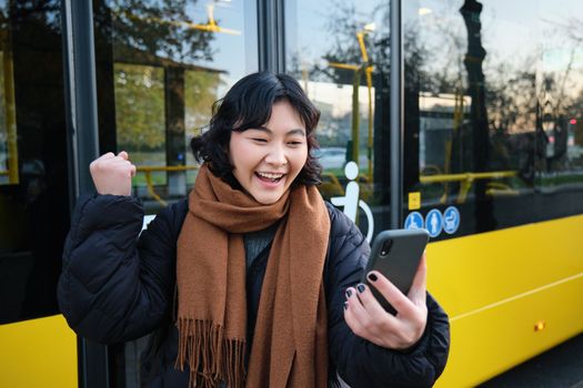 Portrait of cheerful asian girl talks on mobile phone, video chats, looks amazed at smartphone camera, stands on bus stop.