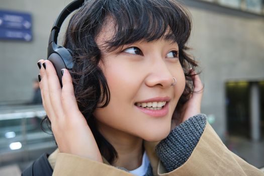 Close up portrait of stylish korean girl standing on street, listening music in headphones and smiling, posing in city centre.