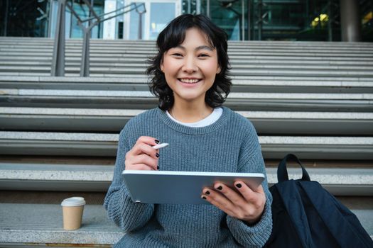 Portrait of asian girl student, hipster sitting on stairs with digital tablet and cup of coffee, draws digital art, makes design project.