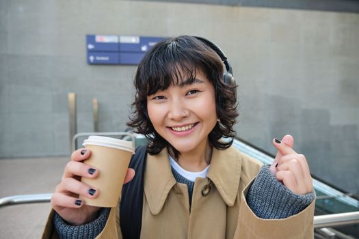 Portrait of stylish asian woman in headphones, drinks coffee to go and smiles, enjoys cappuccino while commutes, stands on street.