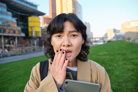 Close up portrait of girl with tablet, looks worried, concerned face expression, feels sad for someone, hears bad news, stands on street near university campus.