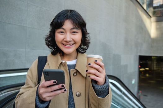 Portrait of hipster girl on escalator, drinking coffee and looking at smartphone, going to work, commute in city, smiling at camera.