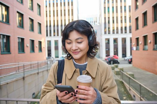 Young woman, tourist in headphones looks at smartphone, drinks coffee to go, checks mobile phone app, listens music and travels. Copy space