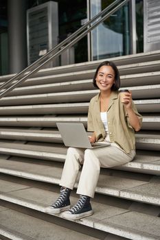 Vertical shot of smiling girl student, asian woman sits on stairs of university campus and drinks coffee, does her homework on laptop.